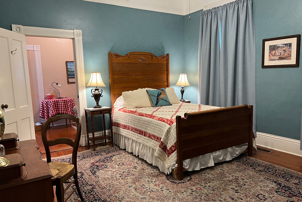 A bedroom with a wooden bed frame, blue walls, and a patterned rug, looking through an open doorway into a room with a red-and-white checkered tablecloth at these San Antonio B&B accommodations.