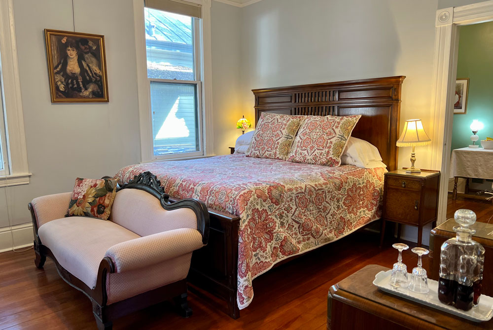 A queen-sized bed with a wooden headboard and floral bedspread in a San Antonio B&B, featuring a small pink upholstered bench at the foot of the bed and a crystal decanter with glasses on a side table.