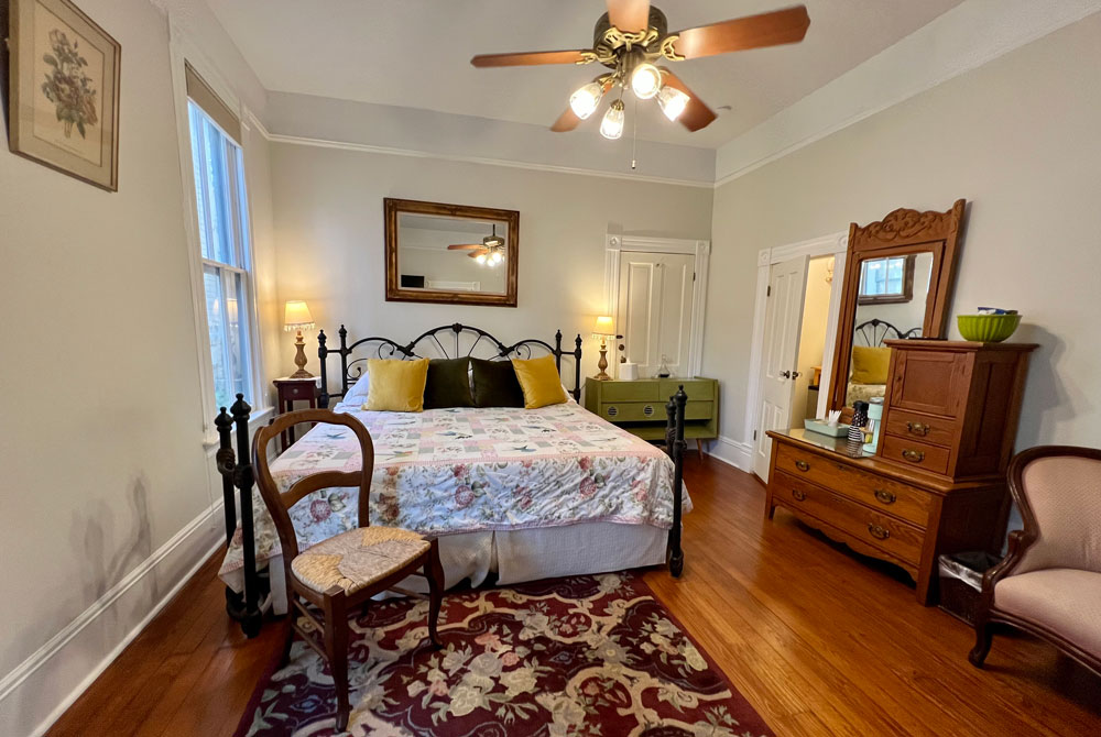 A bedroom with a king-sized bed featuring a floral quilt and yellow pillows, an antique wooden dresser with a mirror, and a patterned area rug on hardwood floors, typical of San Antonio B&B accommodations.