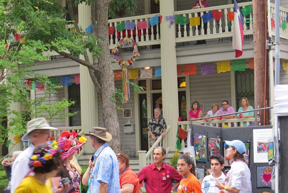 People gather at a two-story house adorned with papel picado banners, a floral wreath, and a Texas flag, marking one of the upcoming events in San Antonio.