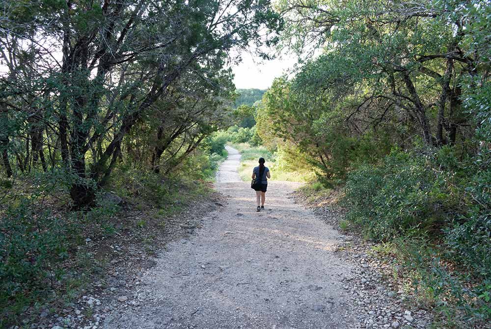 A woman walks away down a gravel path lined by trees, a scenic route for birding in San Antonio.