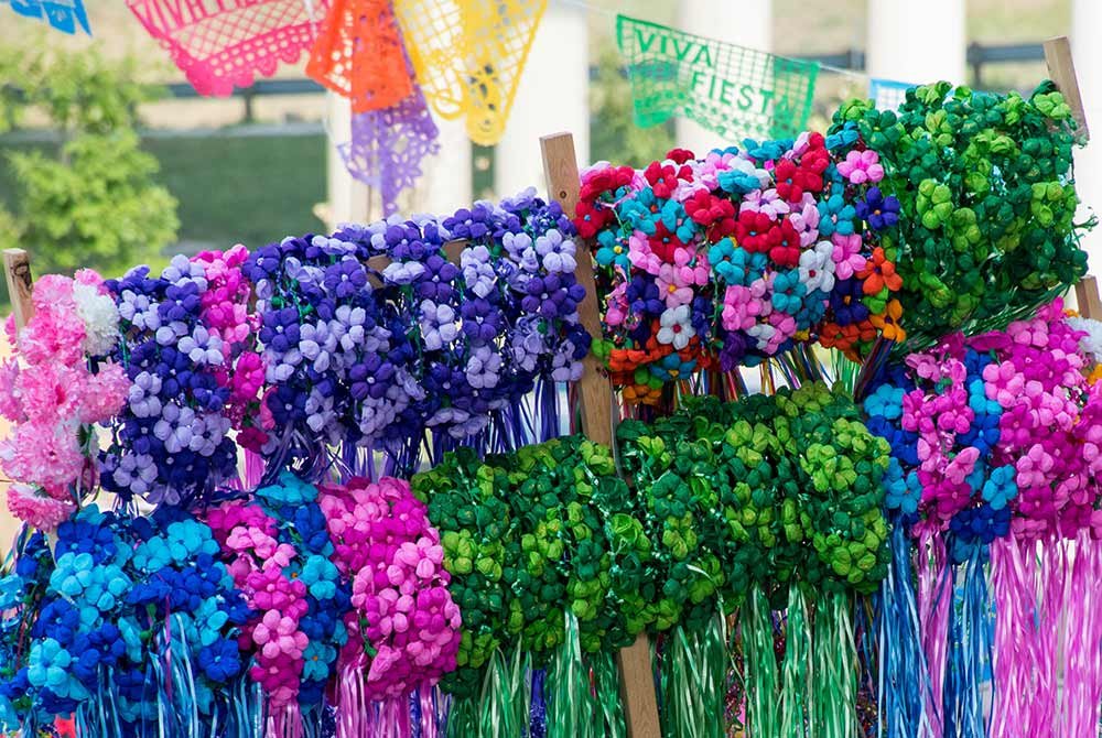Rows of colorful flower crowns with hanging ribbons displayed on a wooden rack, with festive paper banners in the background for upcoming events in San Antonio.