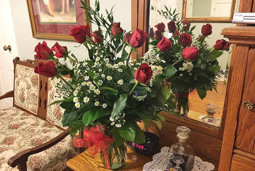 A glass vase filled with red roses and white filler flowers sits on a wooden vanity table, reminiscent of the elegance found in San Antonio getaway packages, in front of a mirror.