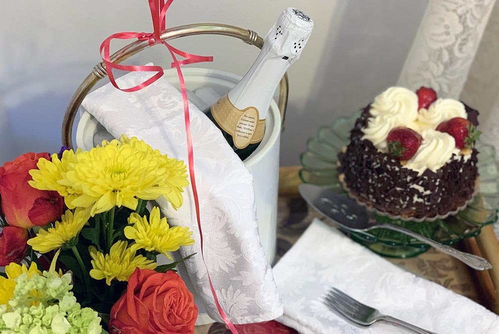 A champagne bottle in a cooler, a bouquet of yellow and red flowers, and a small chocolate cake topped with cream and strawberries on a table for San Antonio getaway packages.