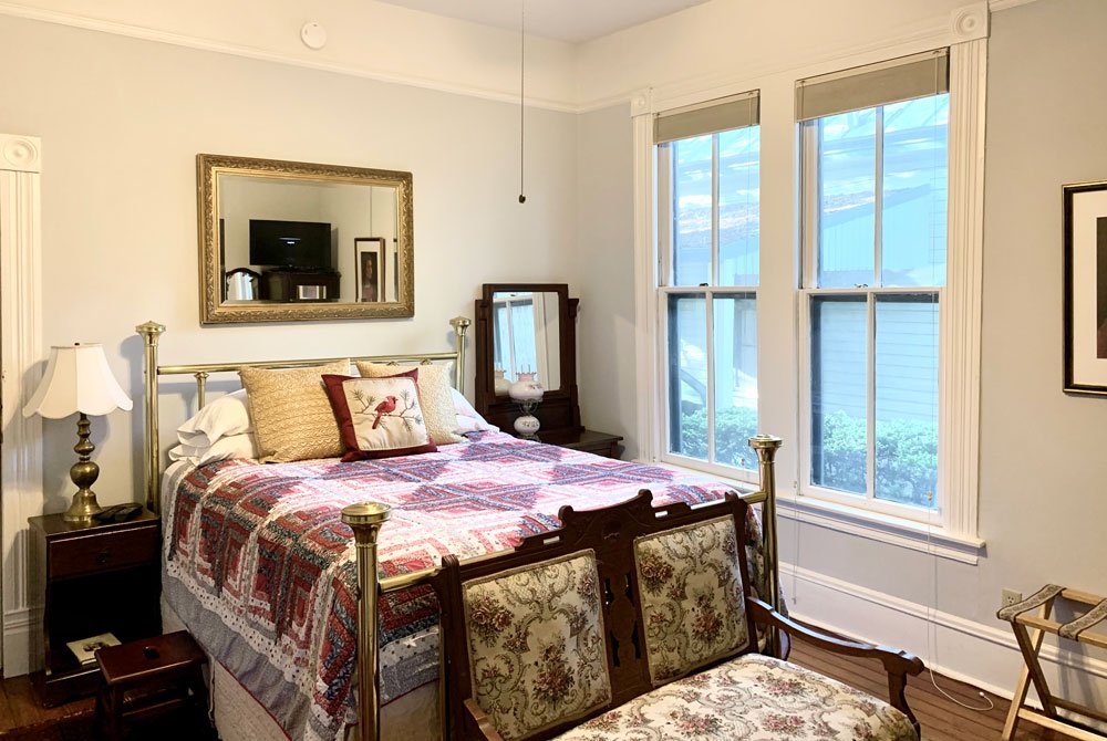 A bedroom featuring a brass-frame bed, a patchwork quilt, and a decorative bench, with a mirror above the headboard and two windows on the wall, perfect for San Antonio B&B accommodations.