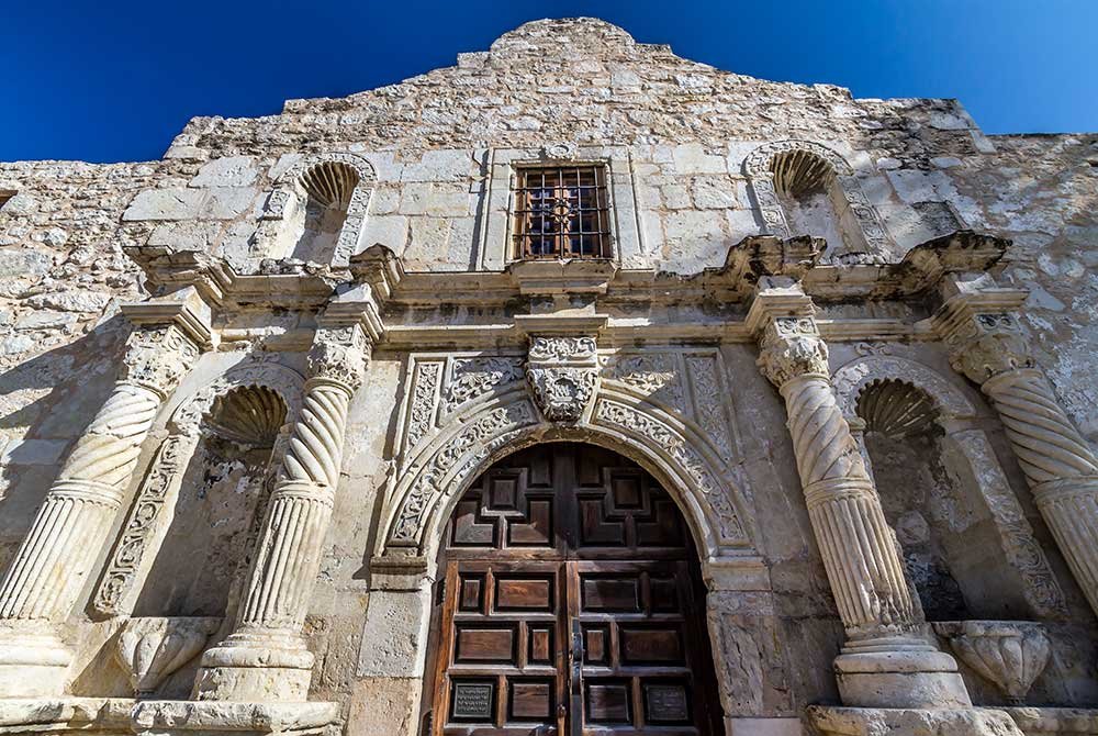 Low-angle view of the Alamo mission facade in San Antonio, Texas, featuring ornate stone columns and a large wooden arched doorway.