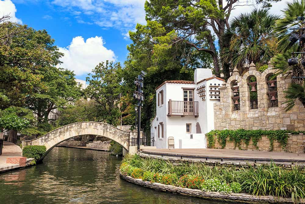 A stone bridge arches over the San Antonio River Walk next to a historic building with arched bell towers, a popular site among things to do in San Antonio, Texas.