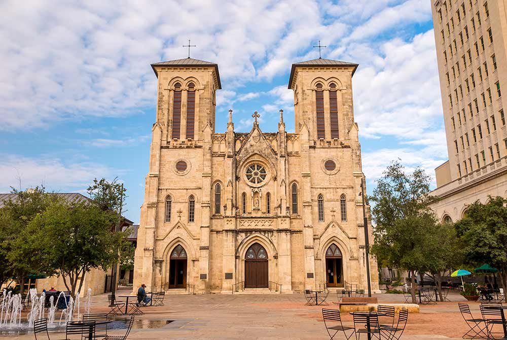 San Fernando Cathedral in San Antonio with two bell towers, seen from a stone plaza with a fountain and cafe tables, a popular highlight of things to do in San Antonio Texas.