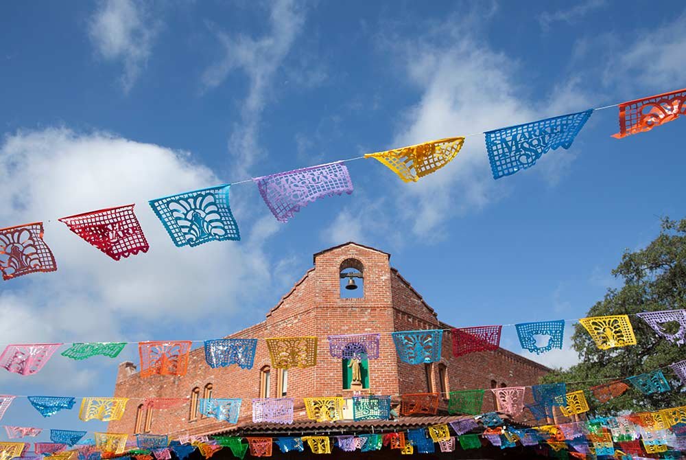 Colorful papel picado banners are strung in front of a historic brick church building in San Antonio, Texas, under a bright blue sky.
