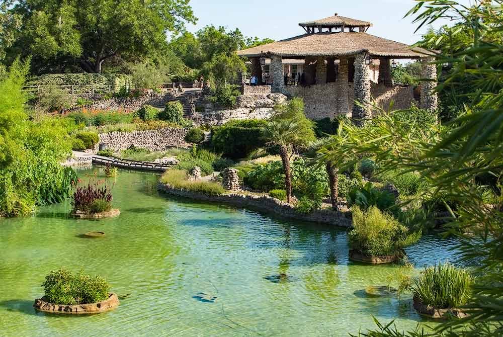 A stone pavilion overlooks a lush, green pond with small islands and a wooden footbridge in a Japanese-inspired garden, a serene highlight among the many things to do in San Antonio, Texas.