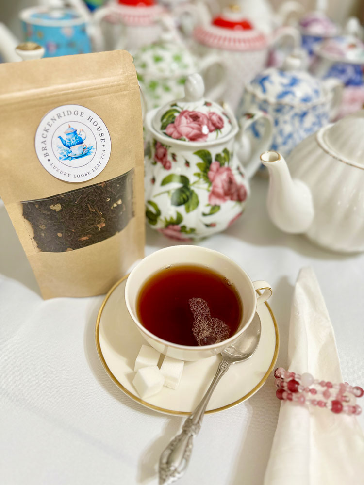 A bag of Brackenridge House Tea Collection loose leaf tea next to a teacup filled with dark tea, with several decorative teapots in the background.
