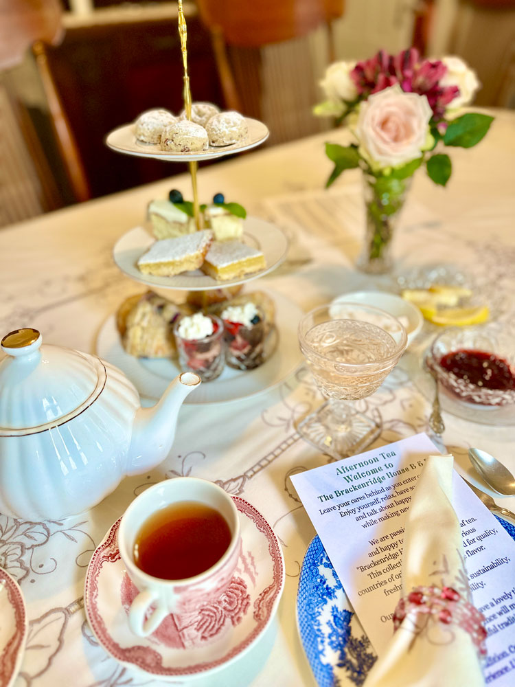 Afternoon tea service from the Brackenridge House Tea Collection, featuring a tiered tray of pastries, a teapot, a cup of tea, and a welcome menu on a table set with floral accents.