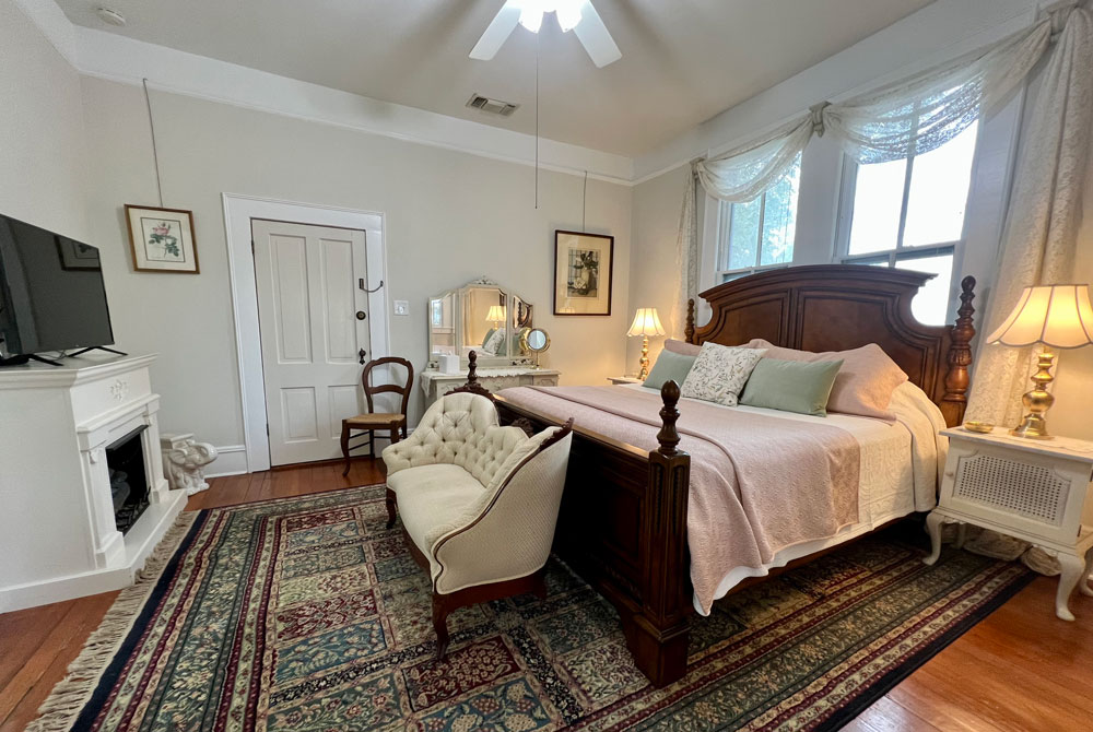A bedroom featuring a large wooden bed with pink bedding, a tufted settee, a white fireplace with a television, and two windows with sheer curtains, representing one of our cozy San Antonio B&B accommodations.